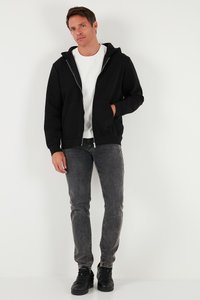 Crna hoodie s patent zatvaračem, prednjim džepovima, u kombinaciji s tamno sivim trapericama i crnim tenisicama. Hoodie ima teksturiranu površinu i srebrni patentni zatvarač.