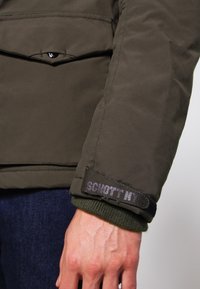 Manche de veste verte avec une sangle noire étiquetée "SCHOTT NY", portée sur un poignet en maille verte et un jean bleu.
