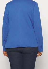 Niebieska marynarka o gładkiej fakturze, zapinana na jeden guzik, z długimi rękawami i ozdobnymi guzikami na mankietach. Widoczne ciemnoniebieskie spodnie poniżej.