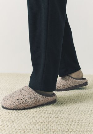 Nohy v béžových chlpatých papučiach a tmavých ponožkách, stojace na textúrovanom svetlom koberci s čiernymi nohavicami.