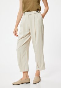 Pantaloni beige dalla vestibilità morbida con pieghe e tasche frontali; presentano un vita alta e un dettaglio con bottoni; tessuto liscio con una leggera lucentezza.