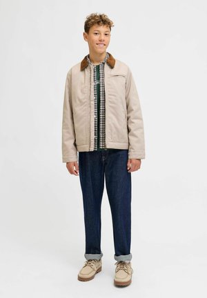 Chaqueta acolchada beige con un cuello de pana marrón, combinada con una camisa de cuadros verde y blanca, pantalones vaqueros oscuros y zapatillas claras.