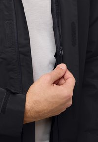 Veste imperméable noire avec fermeture éclair à l'avant et poignets accentués de velcro. Une main est montrée en train d'ajuster la fermeture éclair. La couche intérieure est grise.