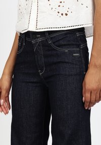 Vaqueros de mezclilla azul oscuro con un corte clásico, que presentan costuras en contraste y una pequeña etiqueta de marca negra. Tejido ligeramente texturizado, cintura de altura media.