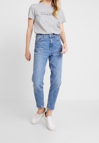 Camiseta de algodón gris claro con logo impreso, combinada con jeans de mezclilla azul de tiro alto. Zapatillas blancas completan el atuendo casual.
