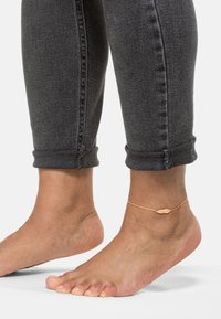 Guldtonet ankelkæde med et muslingedesign, båret på en bar fod. Personen har tætsiddende, mørk grå jeans på, der er rullet op ved anklerne.