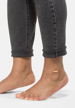 Goldfarbene Fußkette mit Muschel-Design, getragen an einem bloßen Fuß. Die Person trägt enganliegende, dunkelgraue Jeans, die an den Säumen hochgekrempelt sind.