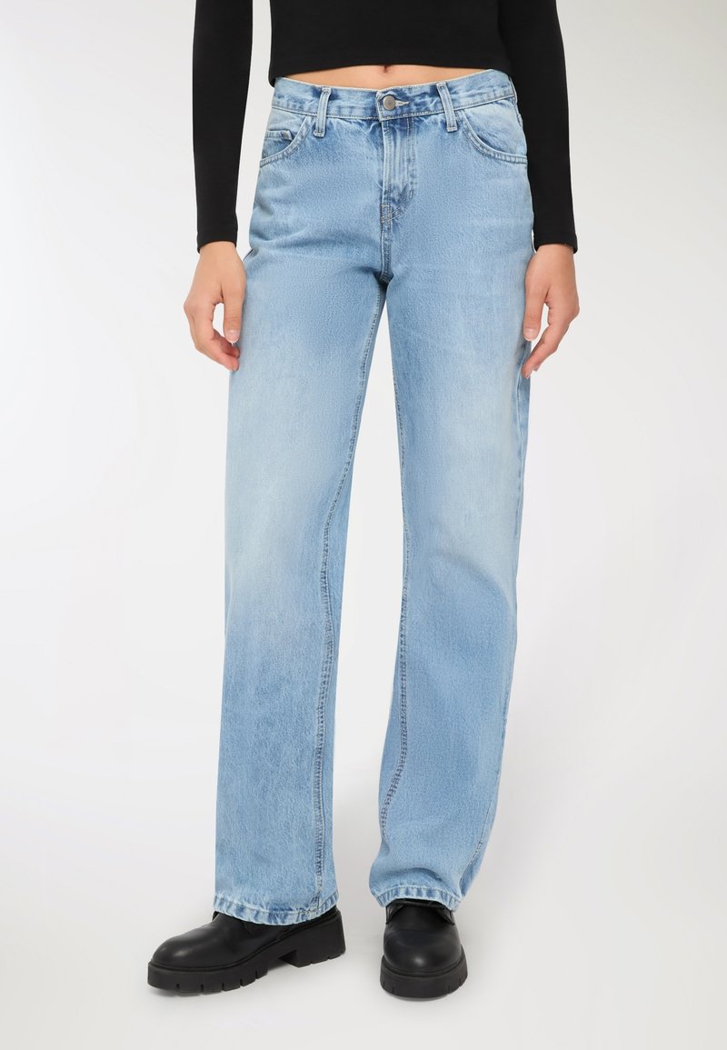 Jeans blu chiaro a vita alta con taglio dritto, caratterizzati da un leggero sbiadimento, passanti per cintura e cinque tasche. Indossati con stivali neri robusti.