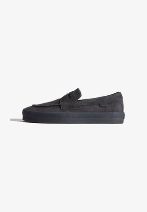 Mocassin noir en daim sans lacets avec une semelle épaisse en caoutchouc et une large bride sur le dessus, présenté de profil sur fond blanc.