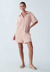 Obleka z kratkimi rokavi svetlo roza barve s krilcem, zapiranjem z gumbi spredaj, enim žepom na prsih in sproščenim krojem. Sledeniče barve ravne sandale.