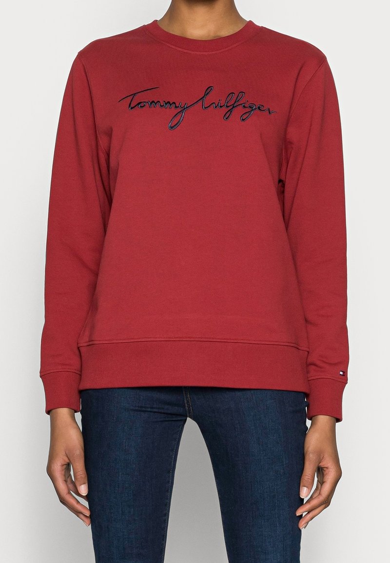 Sweat-shirt bordeaux à manches longues avec broderie "Tommy Hilfiger", porté avec un jean bleu foncé, visible du cou jusqu'aux hanches.