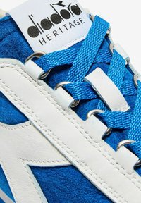 Sneaker blu in suede con dettagli in pelle bianca, caratterizzate da lacci blu testurizzati e occhielli in metallo. Logo Diadora visibile sulla linguetta.