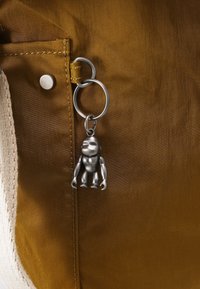Petit porte-clés gorille en métal attaché à un anneau argenté sur un tissu de couleur moutarde avec une poche et des coutures apparentes.