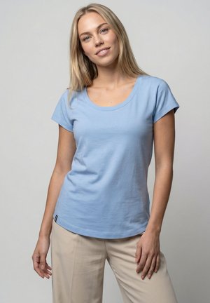 Young woman with long blonde hair wearing a light blue short-sleeve shirt and beige pants, standing against a plain gray background.