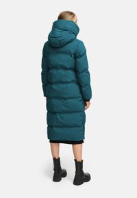 Zeleni puhovski plašč z visokim ovratnikom in kapuco. Ima dolgo dolžino, prešito zasnovo, stranske zadrge in črne poudarke na manšetah.