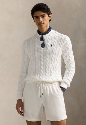 Pull en tricot blanc avec un col rond, associé à un short en lin blanc. Les accessoires comprennent des lunettes de soleil et une montre bleue.