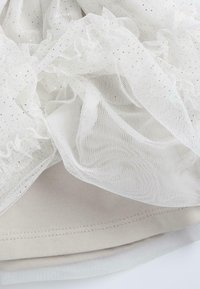 Tulle blanc superposé avec des paillettes dorées, présentant un design froncé, sur un tissu beige lisse. Texture douce avec un ourlet cousu.