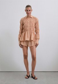 Camisa de color durazno, con botones y un dobladillo peplum; presenta textura fruncida y mangas largas. Combinada con pantalones cortos a juego y sandalias planas.