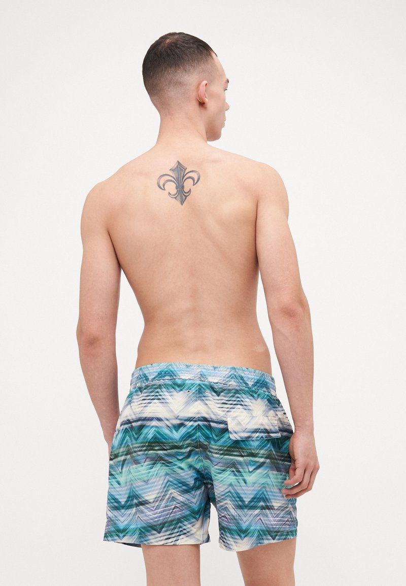 Swim trunks in blue and green wave pattern, elastic waistband, lightweight fabric, side pocket, and two back grommets for drainage.