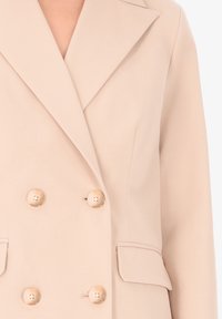 Blazer doppiopetto beige realizzato in tessuto liscio, con grandi bottoni in legno, rever a scollo e tasche laterali.