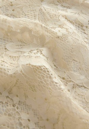 Bēša mežģīņu audums ar ziedu rakstu, raksturots ar smalkām tekstūrām, sarežģītiem detaļām un kopējo mīkstu izskatu.