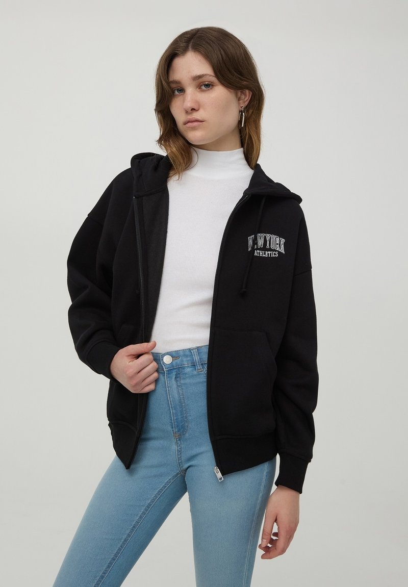 Felpa nera con zip, dotata di tasca frontale e logo "New York Athletics". Realizzata in un tessuto morbido, presenta un fit rilassato e un cappuccio regolabile.