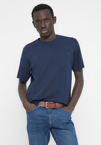 Μπλε ναυτικό κοντομάνικο μπλουζάκι με στρογγυλή λαιμόκοψη και λεπτομέρεια τσέπης, συνδυασμένο με μπλε τζιν παντελόνι και καφέ ζώνη. Λεία υφή υφάσματος.