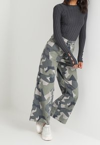 Haut gris à manches longues côtelé associé à un pantalon large camouflage avec des motifs géométriques dans des tons de vert, gris et blanc. Baskets blanches.