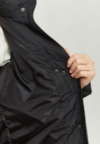 Black padded jacket featuring a smooth exterior, side pockets, and a snap closure. The interior is quilted for added warmth.