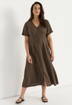 Kobieta ubrana w brązową sukienkę midi z krótkim rękawem i guzikami z przodu oraz czarne sandały na paski, stojąca na jednolitym białym tle.