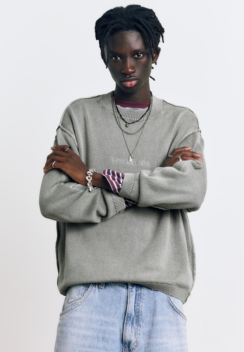 Jeune personne à la peau foncée et aux dreadlocks portant des colliers superposés, un pull gris par-dessus une chemise rayée, et un jean bleu clair, les bras croisés.