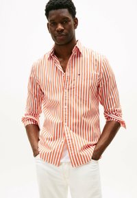 Camicia a righe arancione e bianca, con taschino sul petto e maniche arrotolate, abbinata a pantaloni bianchi.