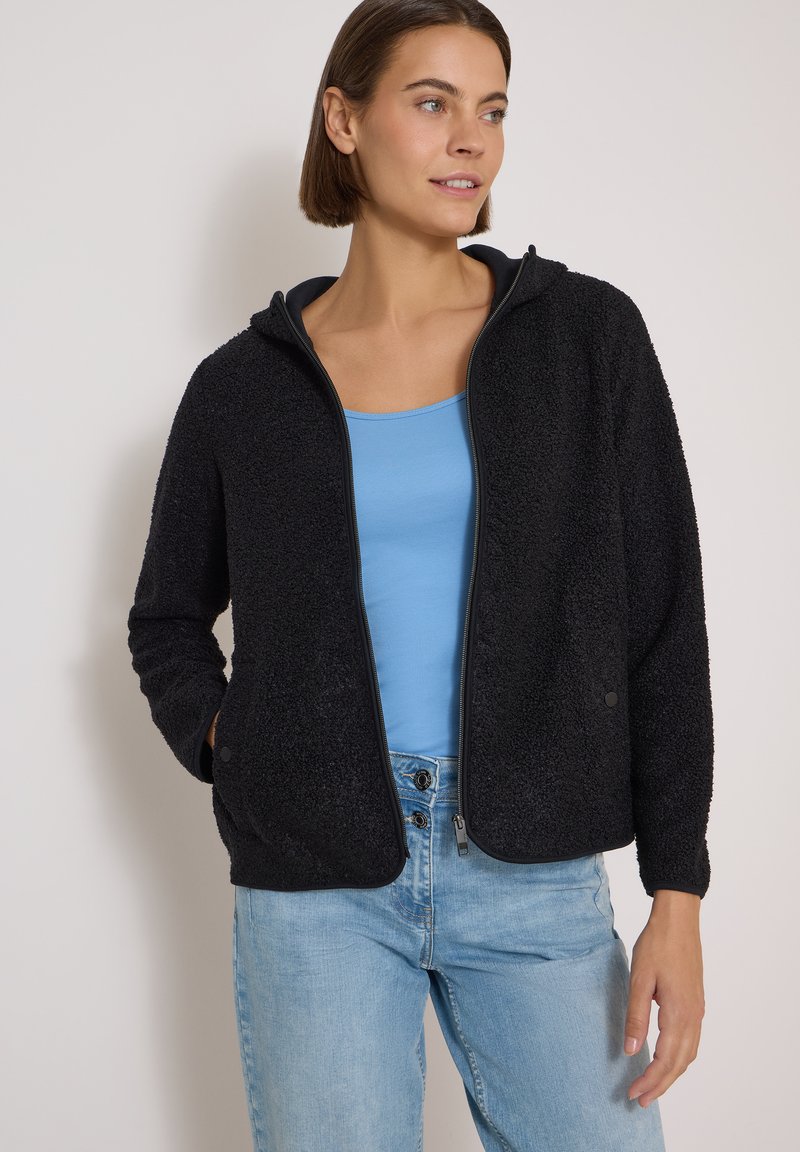 Femme portant une veste polaire noire zippée par-dessus un débardeur bleu et un jean bleu clair, debout contre un mur uni.