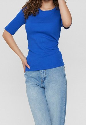 Królewsko niebieska dopasowana koszulka z krótkimi rękawami i okrągłym dekoltem, zestawiona z jasnoniebieskimi dżinsami. Gładka, elastyczna tkanina.