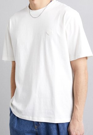 Weißes kurzärmeliges T-Shirt aus Baumwolle mit kleinem Diamantlogo auf der Brust, kombiniert mit blauer Jeans.