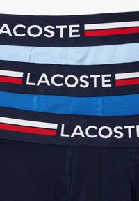 Trois paires de boxers pour hommes avec des ceintures élastiques. Comprend des couleurs marine, bleu clair et bleu foncé ; logo Lacoste affiché en évidence.