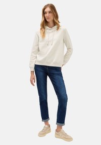 Donna con felpa beige chiara, jeans blu scuro arrotolati e sneakers beige, in piedi con una mano in tasca su sfondo bianco.