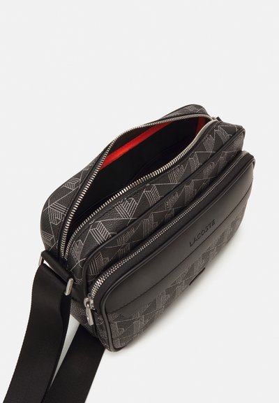 Sac bandoulière noir texturé avec quincaillerie argentée, présentant un motif géométrique, un intérieur rouge et une sangle ajustable. Logo LACOSTE présent.