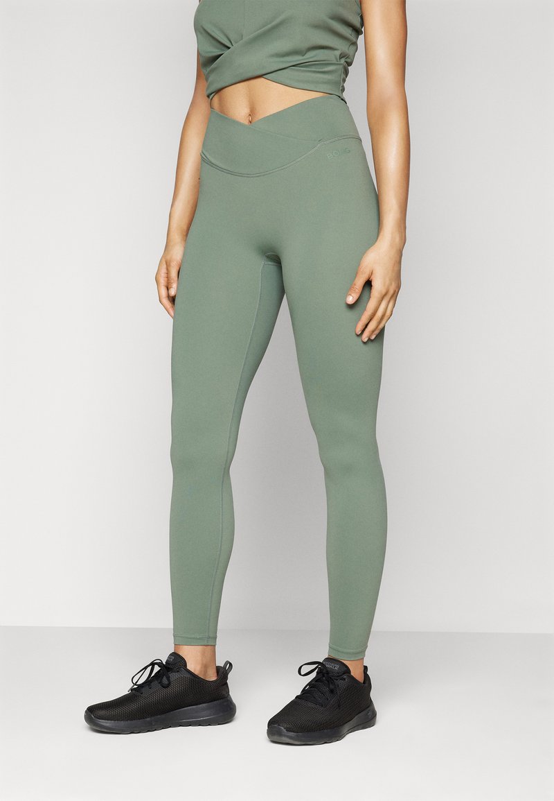 Oliven grønne tights med høy, kryssbelte og flatlock-sømmer, kombinert med svarte treningssko for kontrast.