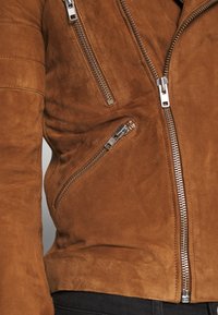 Giacca in suede marrone con molteplici zip in argento, una finitura texturizzata e un design aderente con dettagli trapuntati sulle maniche.