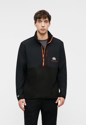 Νέος άνδρας που φοράει μαύρο fleece μπουφάν με πορτοκαλί φερμουάρ και λογότυπο στο στήθος, στέκεται μπροστά σε ένα απλό φωτεινό φόντο.