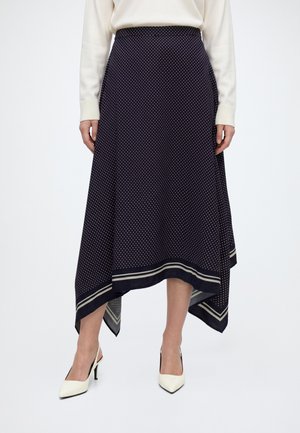 Femme portant une jupe bleu marine à pois avec un ourlet asymétrique et des rayures blanches près du bas, associée à des chaussures blanches à talons hauts.