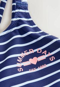 Top de înot dungat de culoare albastru închis, cu dungi de culoare albastru deschis. Prezintă un text imprimat roz "ZILE DE VARĂ" cu un inimi și detaliul "EST. 1982". Material neted.