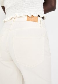 Jean taille haute beige clair avec une texture lisse, comportant deux poches arrière et une étiquette en cuir marron estampillée "jeans parfaits".