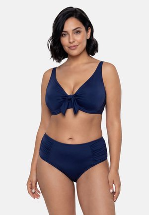 Kvinde med mørkt hår iført en navyblå bikini med en top med snøre foran og rynkede højtaljede underdele, stående mod en lys baggrund.