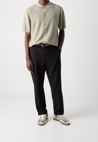 Lichtgroene polo met korte mouwen en een ribbelstructuur, met een knoopsluiting aan de voorkant, gecombineerd met zwarte broek en grijze sneakers met groene en roze accenten.