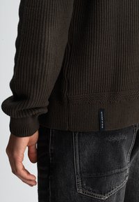 Dunkelbrauner Rippstrickpullover mit einem enganliegenden Bund, strukturiertem Detail und einem kleinen Etikett am Saum. Kombiniert mit dunkler Jeans.