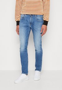 Person wearing light blue jeans, white sneakers, and a long-sleeve orange and white striped shirt, standing against a plain background.