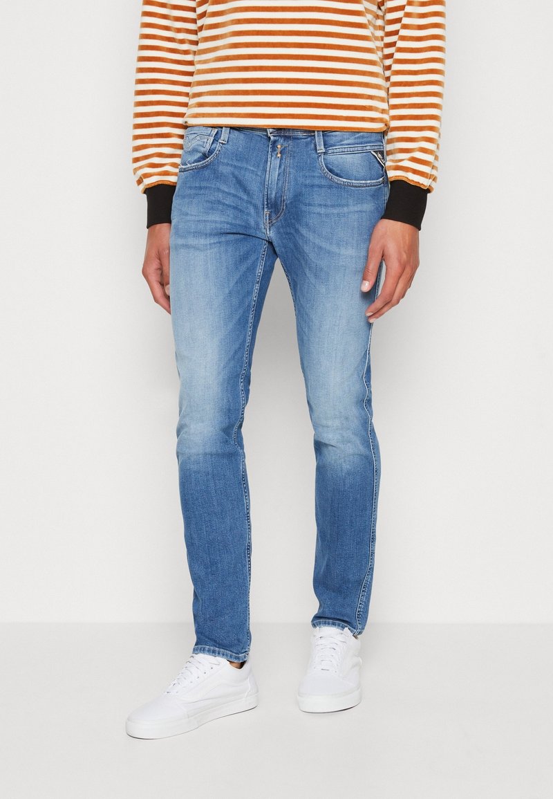 Person wearing light blue jeans, white sneakers, and a long-sleeve orange and white striped shirt, standing against a plain background.