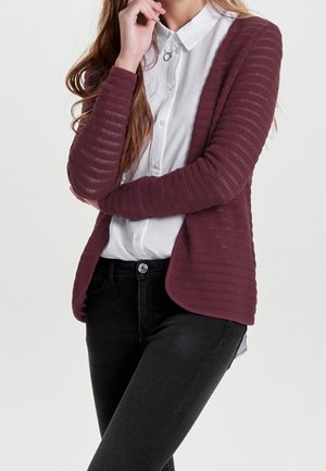 Chaqueta de punto texturizada color burdeos con frente abierto, mangas largas y diseño acanalado, combinada con una camisa blanca con cuello y vaqueros negros.